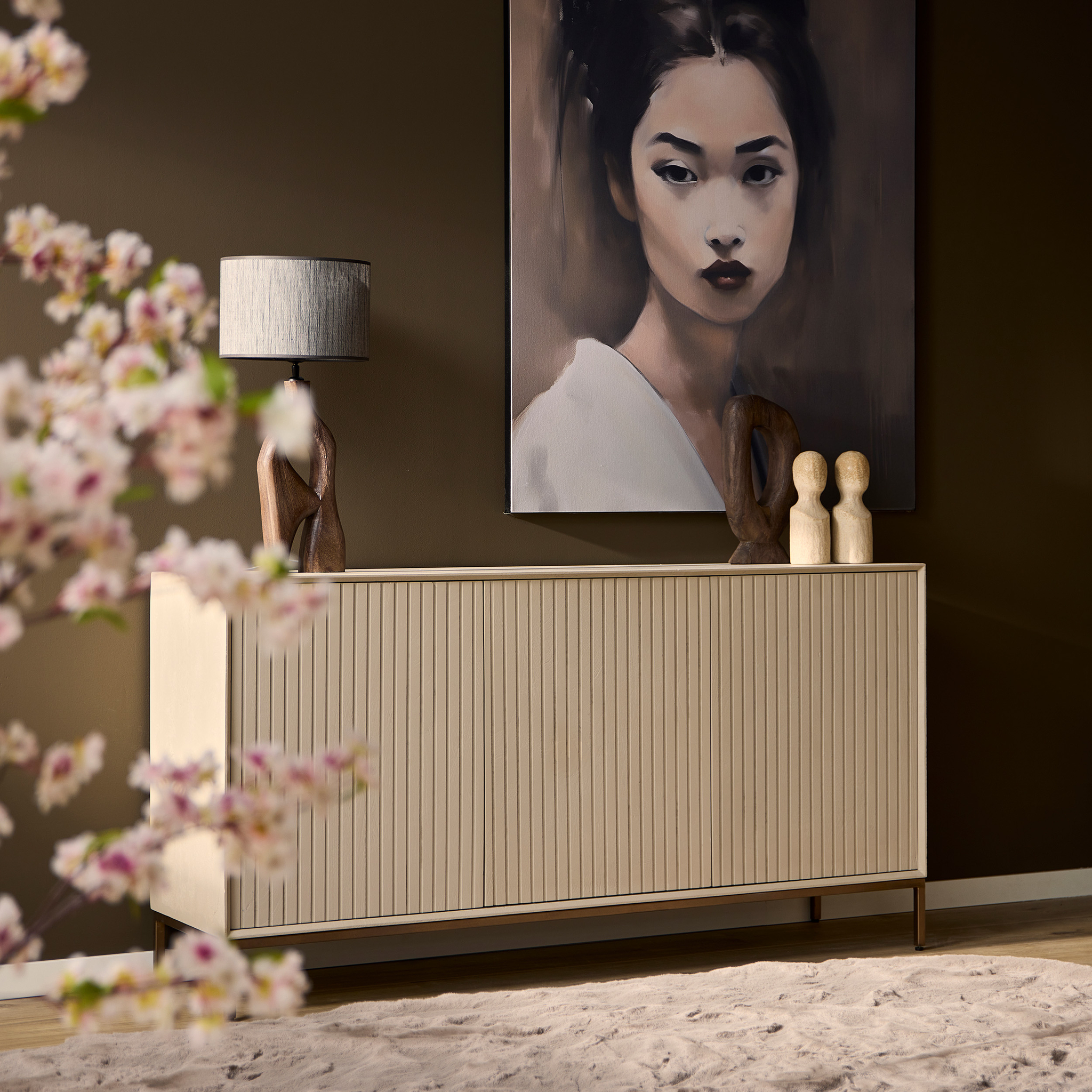 Een minimalistische kamer met de Dressoir Madison Sand 165 cm, abstracte houten sculpturen, een tafellamp en een groot vrouwenportret op een bruine muur. Roze bloemen verschijnen vaag op de voorgrond.