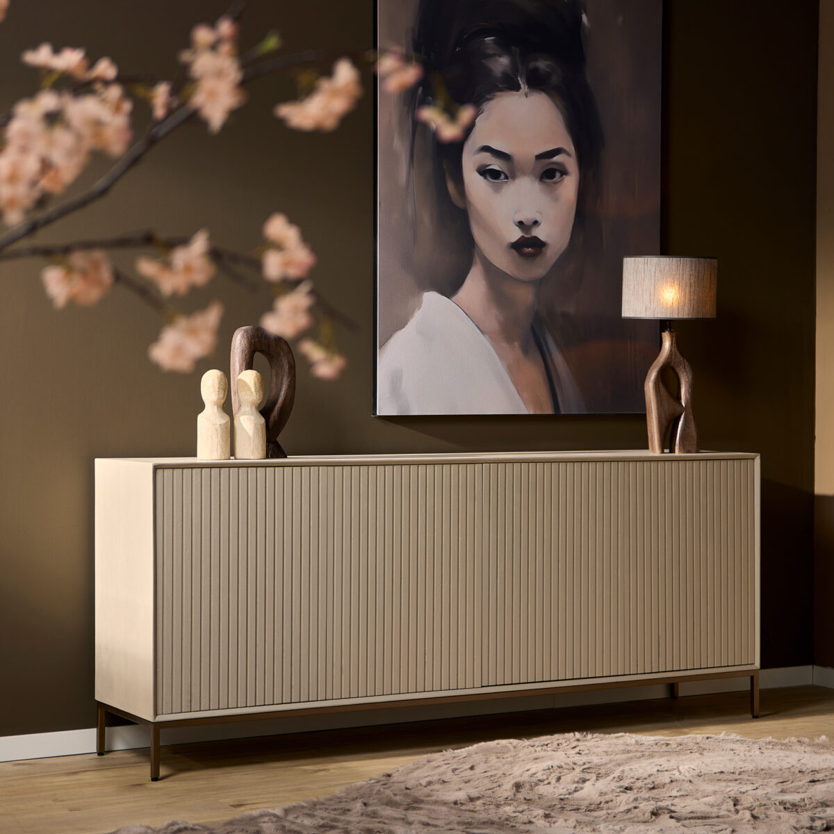 De Dressoir Madison Sand | 210 cm staat tegen een bruine muur met sculpturen en een lamp erop, onder een groot vrouwenportret. Vage kersenbloesemtakken staan op de voorgrond en een tapijt met textuur bedekt een deel van de houten vloer.