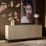 De Dressoir Madison Sand | 210 cm staat tegen een bruine muur met sculpturen en een lamp erop, onder een groot vrouwenportret. Vage kersenbloesemtakken staan op de voorgrond en een tapijt met textuur bedekt een deel van de houten vloer.