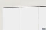 Close-up van het bovenste gedeelte van de Opbergkast 'Ray' 3 deuren en 4 legplanken Wit (2c), een minimalistische witte kast, tegen een lichte muur; een klein Housebad-logo verschijnt in de rechterbenedenhoek.