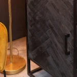 Een close-up van de Dressoir New York | 165 cm met een donker houten visgraatmotief, getoond naast een mosterdgele stoel en een gouden vloerlamp op een grijze vloer.