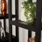 Een close-up van de Boekenkast Boaz Black | 3 laden plank met een witte vaas met groene wingerd, een koperkleurige handsculptuur en kleine decoratieve items.