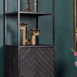 De Boekenkast New York, gemaakt van mangohout en staal met een zwarte deur met chevronpatroon, toont gouden geometrische kaarsenhouders en glazen decor tegen een donkergroene muur. Roze bloemen verschijnen gedeeltelijk aan de rechterkant.