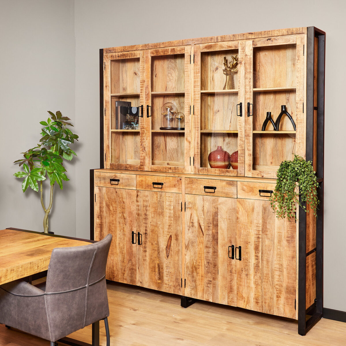 De Vitrinekast Boaz XXL, een houten kast met glazen deuren, toont decoratieve voorwerpen en planten tegen een beige muur naast een bebladerde potplant, met een houten tafel en grijze stoel gedeeltelijk zichtbaar op de voorgrond.