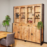De Vitrinekast Boaz XXL, een houten kast met glazen deuren, toont decoratieve voorwerpen en planten tegen een beige muur naast een bebladerde potplant, met een houten tafel en grijze stoel gedeeltelijk zichtbaar op de voorgrond.