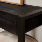 Close-up van de Sidetable Boaz Black | 120 cm, met een zwarte houten hoek met een stevig metalen frame, een glimp van een beige beklede zitting eronder en een object met houten frame dat bovenop rust.