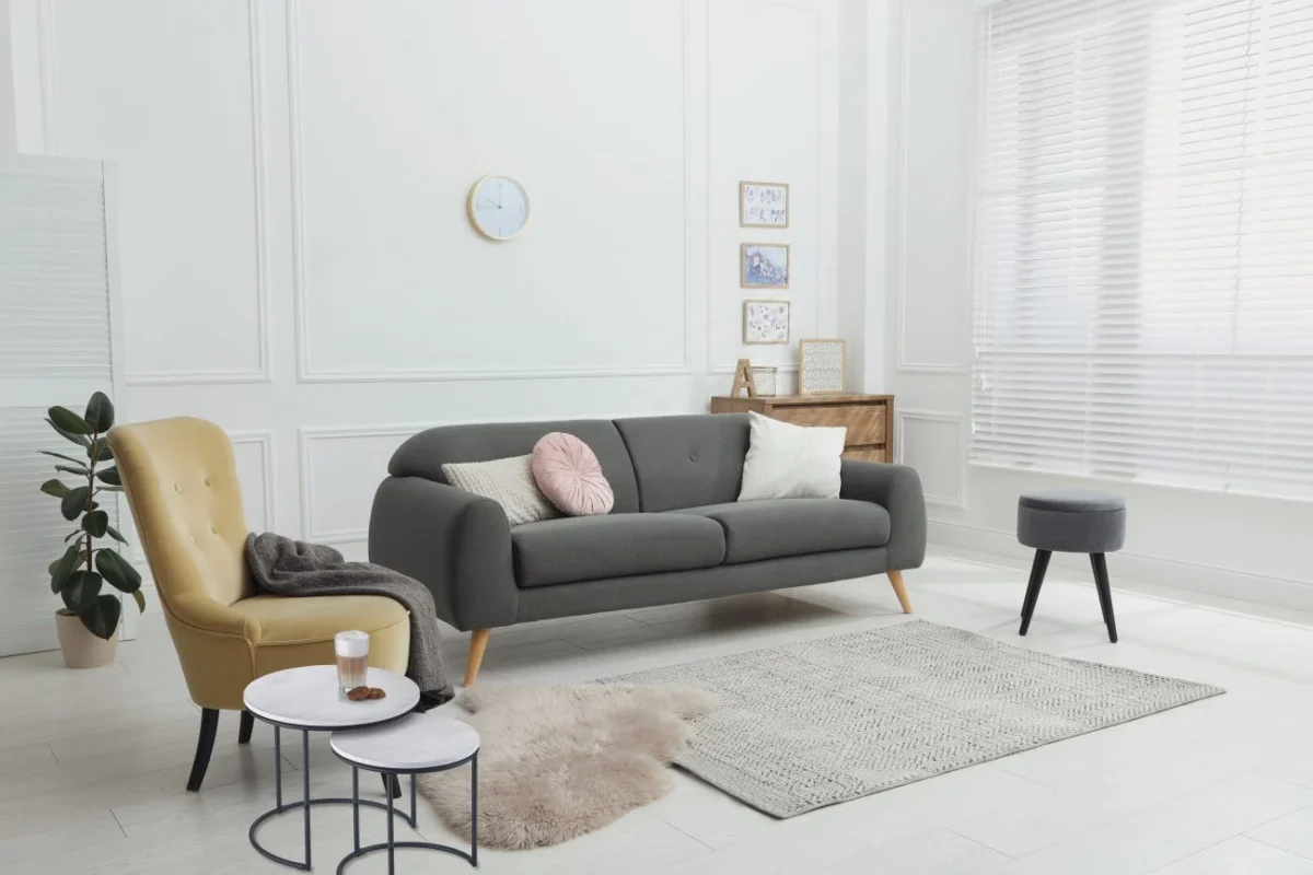 Een moderne woonkamer heeft een grijze bank, een gele fauteuil met een bruin kleed, de Mirrors and More Bijzettafel DOREEN met een kopje, een grijze kruk, een vloerkleed met structuur, een plant en grote ramen met witte jaloezieën die natuurlijk licht binnenlaten.