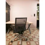 Een Workliving Zuidas Mesh - Regain zitting - Bureaustoel Ergonomisch Ontwerp (N)EN 1335 kijkt uit op een houten bureau in een minimalistische kamer met lichtroze muren, tegelvloer met patroon, pennenhouder, notitieblok en een deur en raam op de achtergrond.