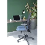 Een moderne werkplek met een laptop, glazen kan, lamp op een houten bureau tegen een groene muur, grote potplant vlakbij en de Workliving Nora Mesh Blauw Regain ergonomische bureaustoel (NEN1335) ervoor.