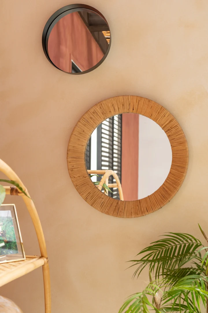 Een kleine ronde spiegel met een zwarte metalen rand hangt aan een beige muur. Op de voorgrond staat een groene bladplant en een deel van een lichte houten plank met een ingelijste foto.