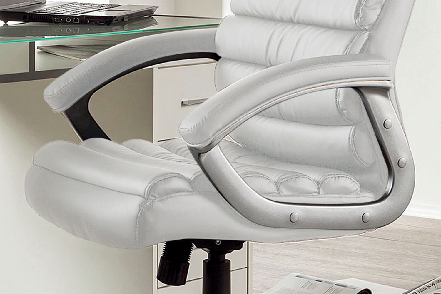 Een close-up van de SALE - Bureaustoel 'Kennedy' PU Wit (1c), een witleren bureaustoel met gecapitonneerde armleuningen, geplaatst naast een glazen bureau met een deel van een laptop en lade in beeld.