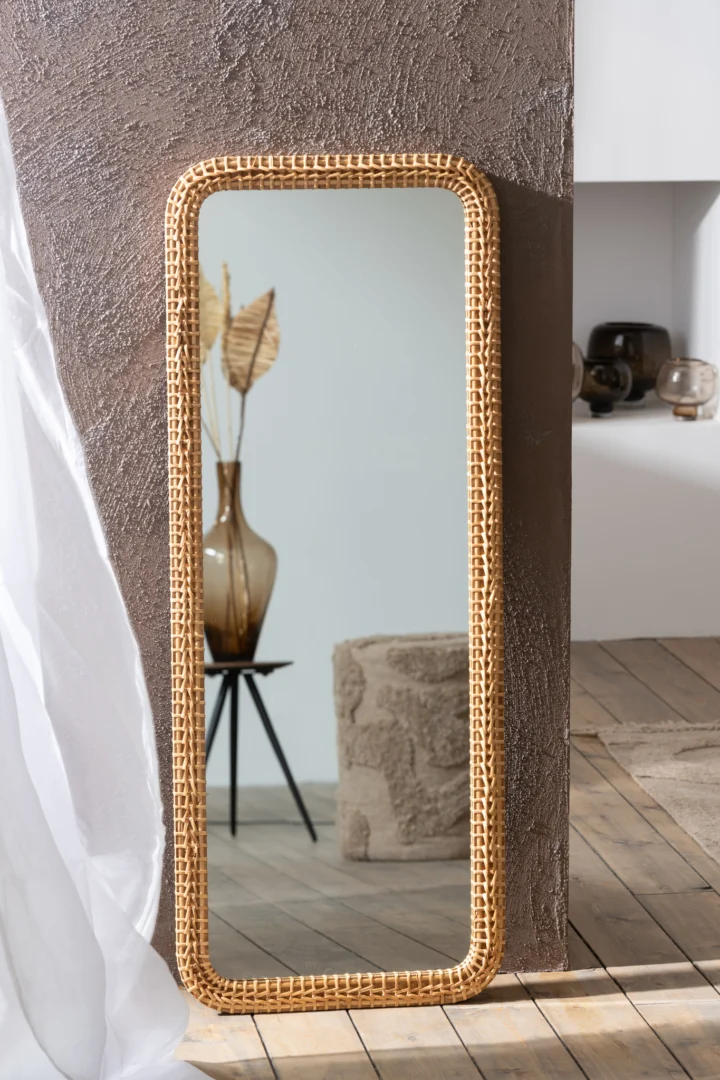 De Mirror Rectangle Fir Wood/Glass Natural leunt tegen een bruine muur met textuur en weerspiegelt een vaas met gedroogde bladeren op een driepoottafel, een beige kruk met textuur en een deel van een moderne kamer met neutrale tinten.
