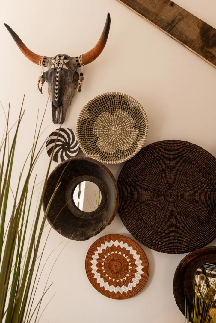 Wanddecoratie met een decoratieve schedel, gevlochten manden en de spiegel Round Bowl Shaped Recycled Wood Brown voegt geometrische en natuurlijke patronen toe boven hoge groene grassen op de voorgrond.