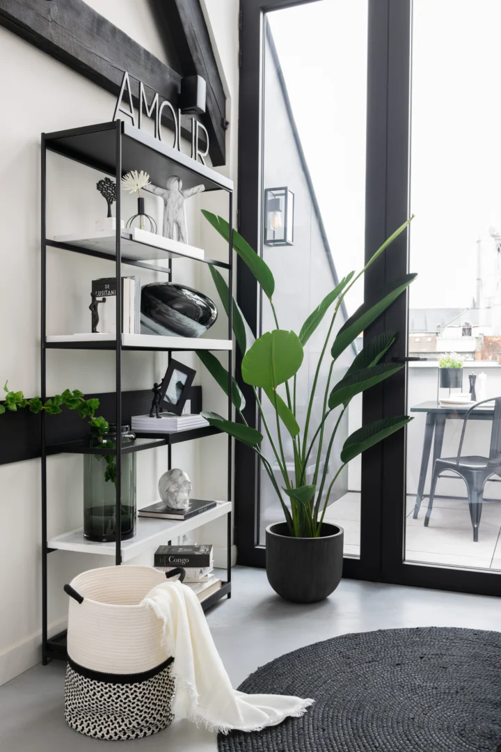 Een moderne woonkamer is voorzien van Vloerkleed Rond Jute Zwart, een zwarte metalen plank met decor, een grote potplant, een deken in een mand en glazen deuren van vloer tot plafond die leiden naar een balkon met zitplaatsen buiten.