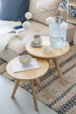 In een gezellige woonkamer staan op twee ronde houten tafels een kaars, mok, snackschaal, glazen vaas met witte pompons en een notitieblok. Het Vloerkleed Denim Katoen Blauw/Wit Groot ligt eronder; een bank met kussen en plaids staat op de achtergrond.
