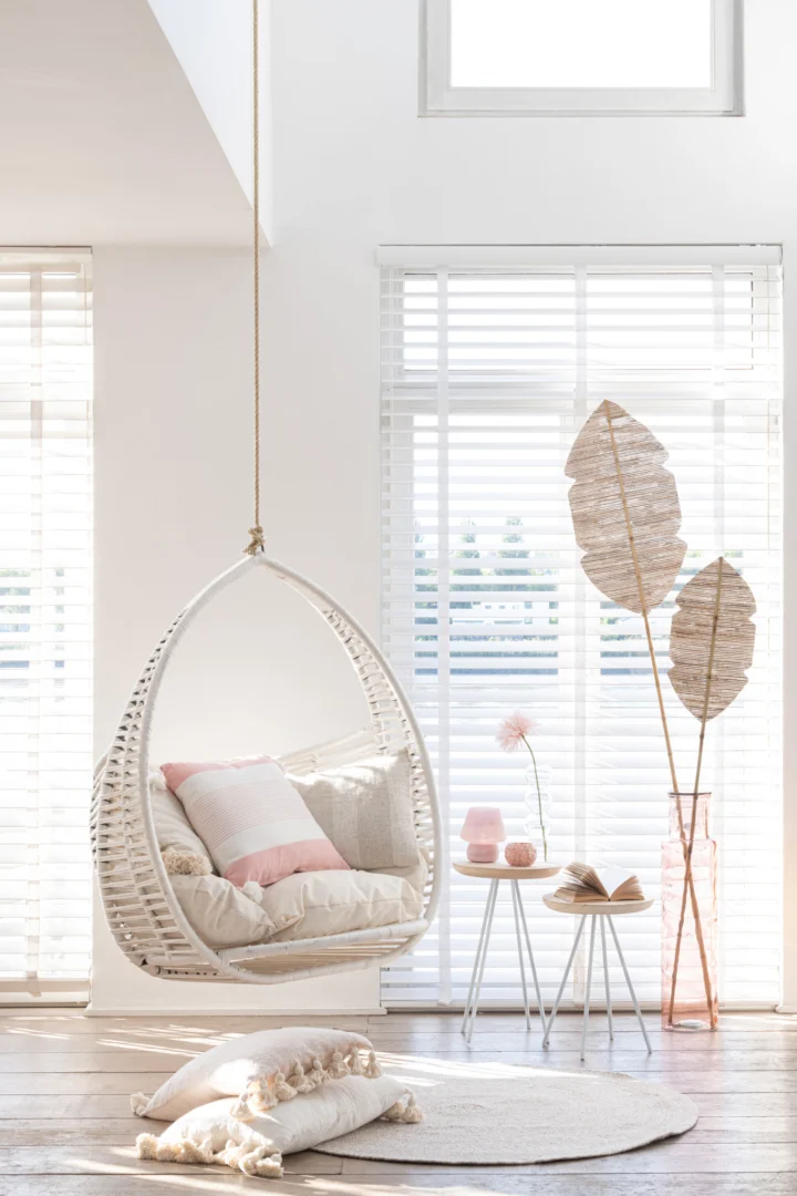Een lichte, luchtige kamer is voorzien van het Vloerkleed Miami Outdoor Poly Naturel/Wit Medium, grote ramen met witte jaloezieën, een rotan stoel gevuld met kussens en een roze kussen, twee witte bijzettafels met boeken en kaarsen, en vloerkussens.