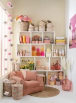 In een gezellige, kleurrijke kamer ligt het Vloerkleed Rond Gevlochten Zeegras Naturel onder een zachte stoel met manden en kussens, bij een witte kubusvormige kast vol met woonaccessoires en een zonovergoten raam versierd met roze en witte pompons.