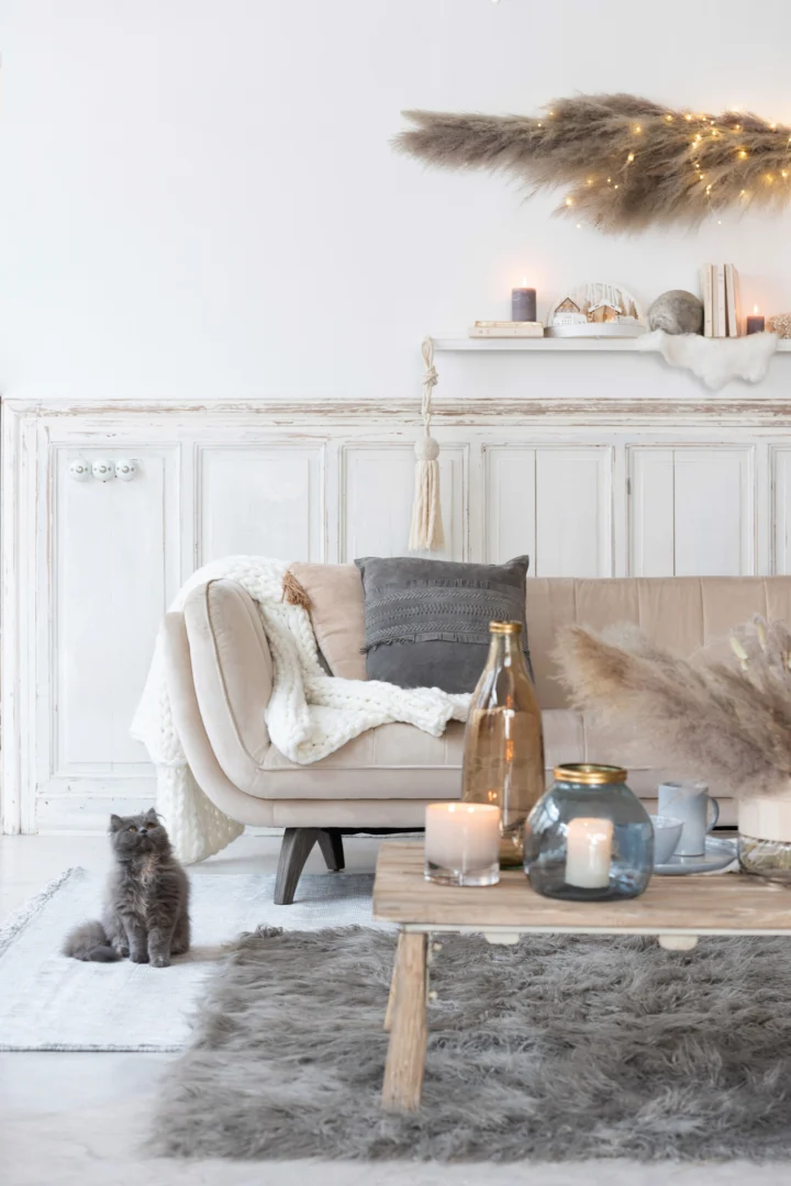 Een gezellige woonkamer met een beige bank, grijze kussens, een gebreide deken, het Vloerkleed Langharig Acryl Grijs vloerkleed, een houten salontafel met kaarsen en glazen flessen, pampagrasdecor en een grijze kat op de vloer.