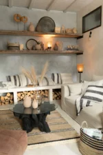 Gezellige woonkamer met neutrale tinten, gestreepte kussens, rustieke houten planken, haardhoutopslag, een glazen salontafel en het Vloerkleed Gevlochten Zeegras-Palmblad Naturel/Zwart vloerkleed voor natuurlijke geweven accenten.