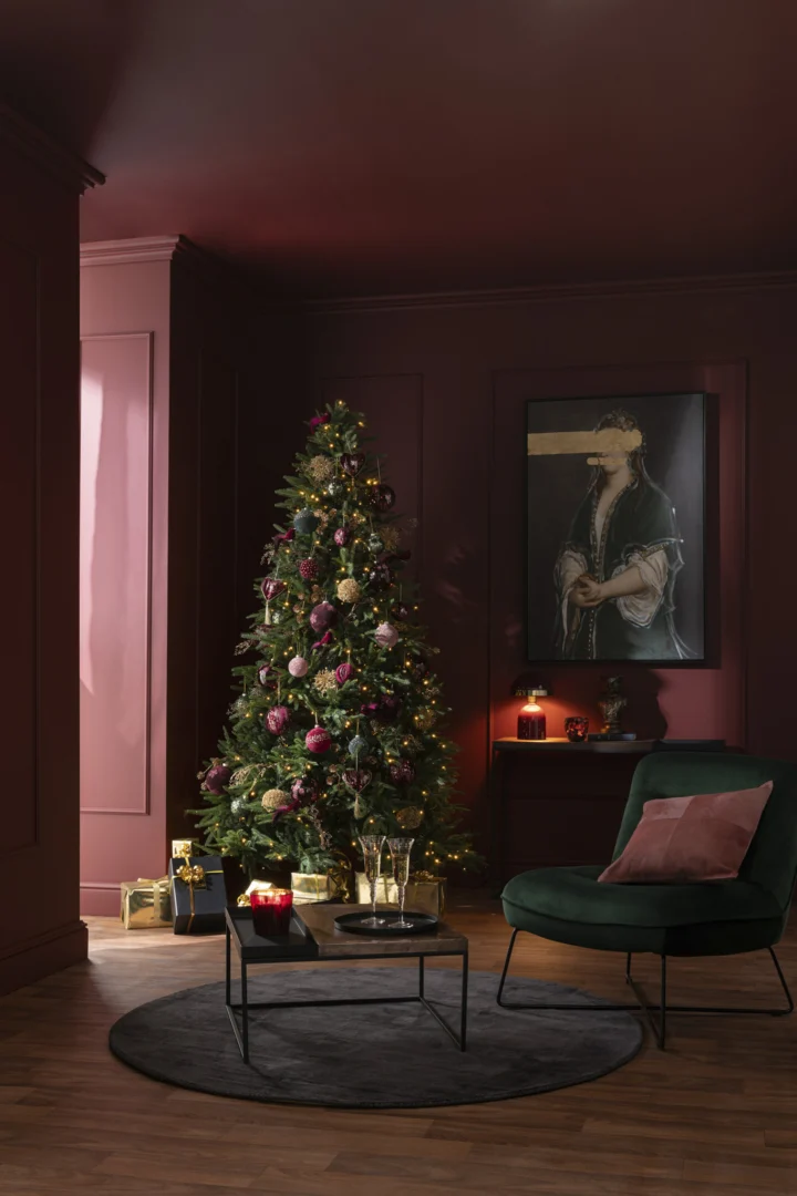 Een versierde kerstboom met roze en gouden ornamenten staat op de Vloerkleed Rond Handgemaakt Viscose Zwart in een donkere woonkamer met bordeauxrode muren, een groene stoel, cadeautjes onder de boom en een ingelijst kunstwerk.