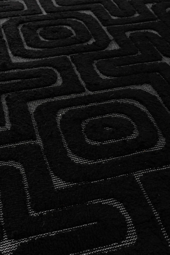 Close-up van Vloerkleed Reliëf Viscose Zwart: zwarte stof met structuur en een geometrisch patroon van vierkanten en concentrische rechthoeken, geaccentueerd met lichtgrijze stippellijnen.