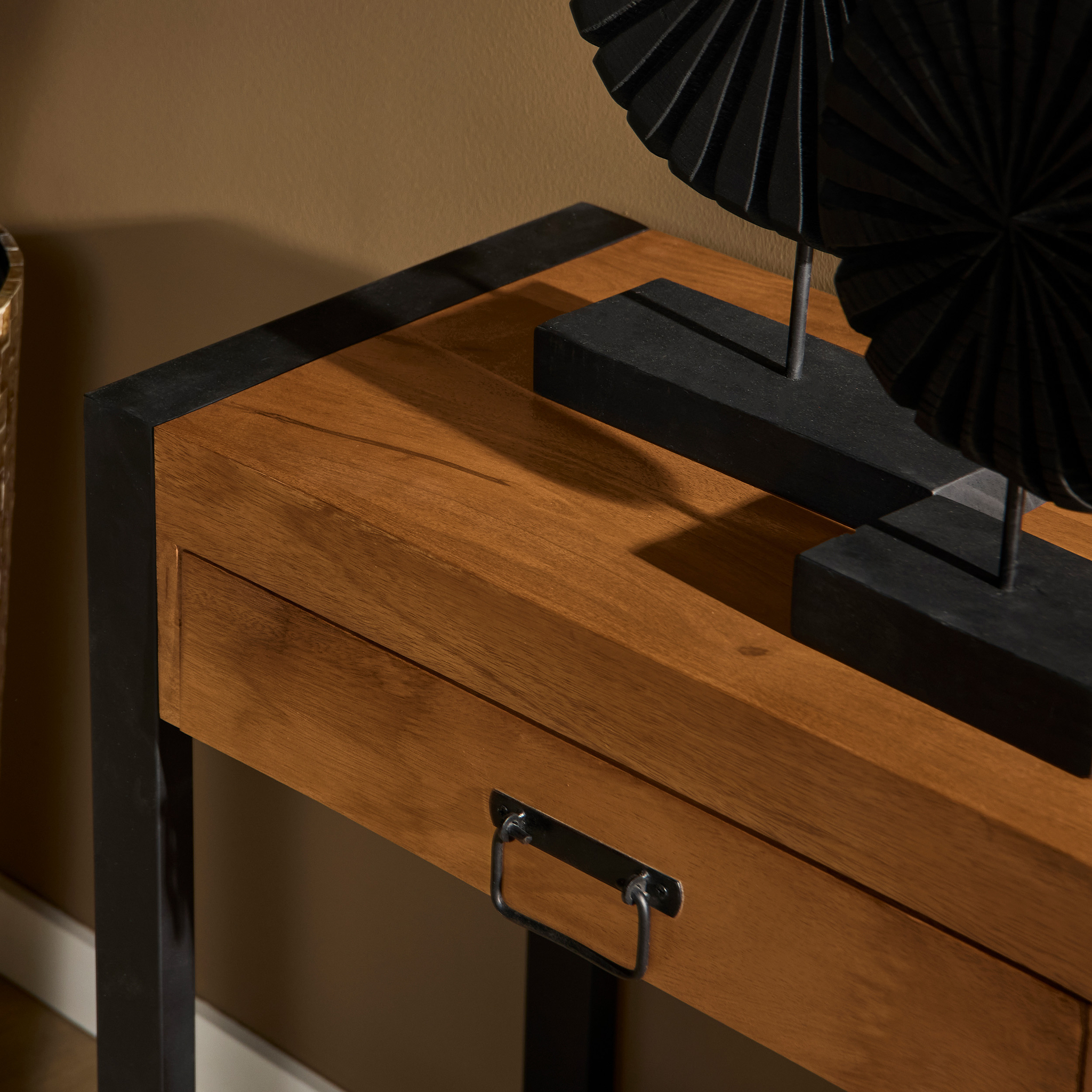 Een close-up van de Sidetable Boaz | 120 cm, een houten consoletafel met zwarte metalen poten en een rechthoekig ladehandvat, afgewerkt met zwart geometrisch decor, tegen een bruine muur.