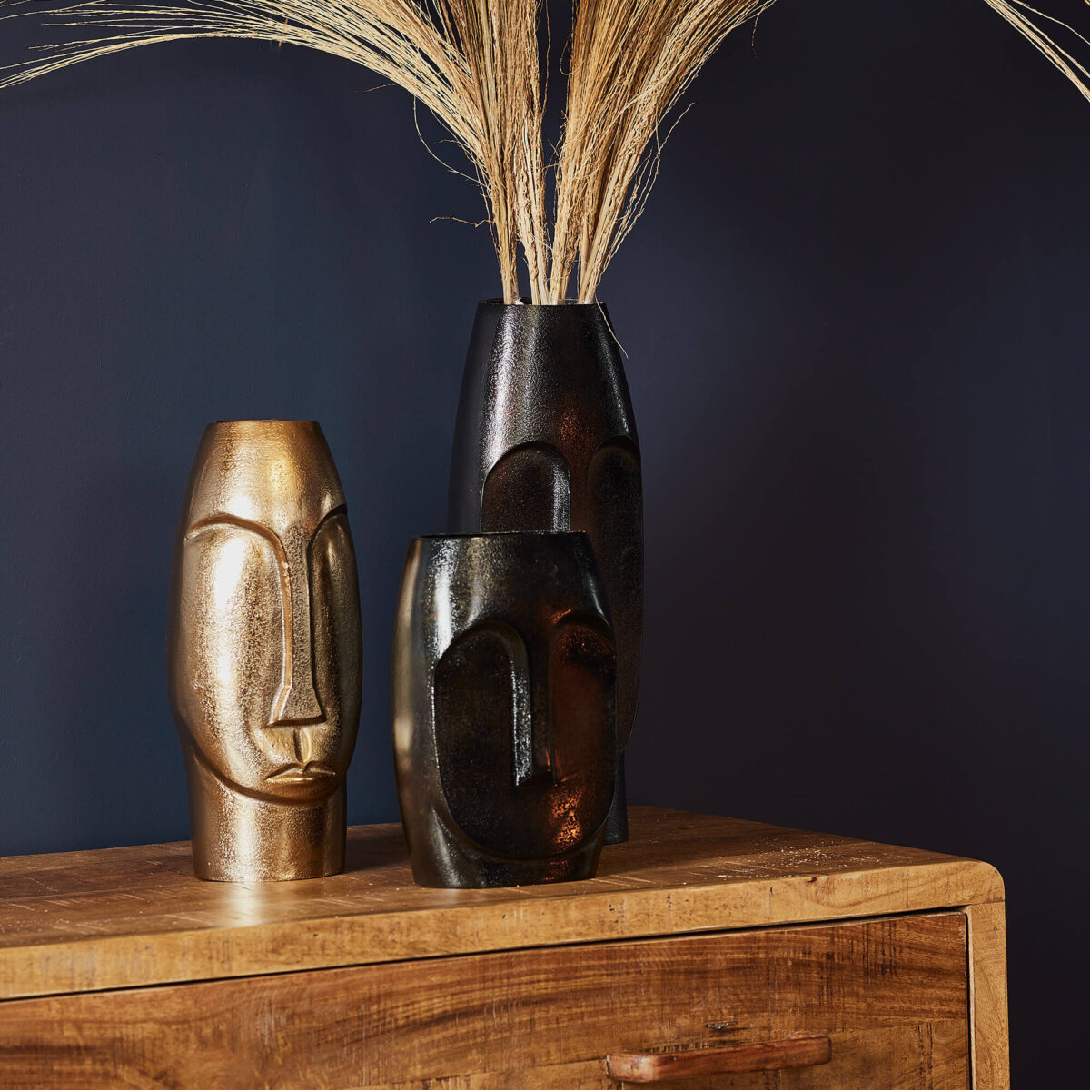 De Vaas Grace - Zwart | Small, een sculpturale zwarte vaas met een abstract gezichtsontwerp, staat op een houten dressoir met gedroogd pampagras tegen een donkerblauwe achtergrond.