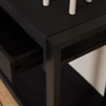 Close-up van de Sidetable Boaz ZWART | 180 cm, een moderne zwarte tafel met slanke metalen randen en een gedeeltelijk open lade. Verschillende witte cilindervormige objecten staan op het tafelblad boven een houten vloer.