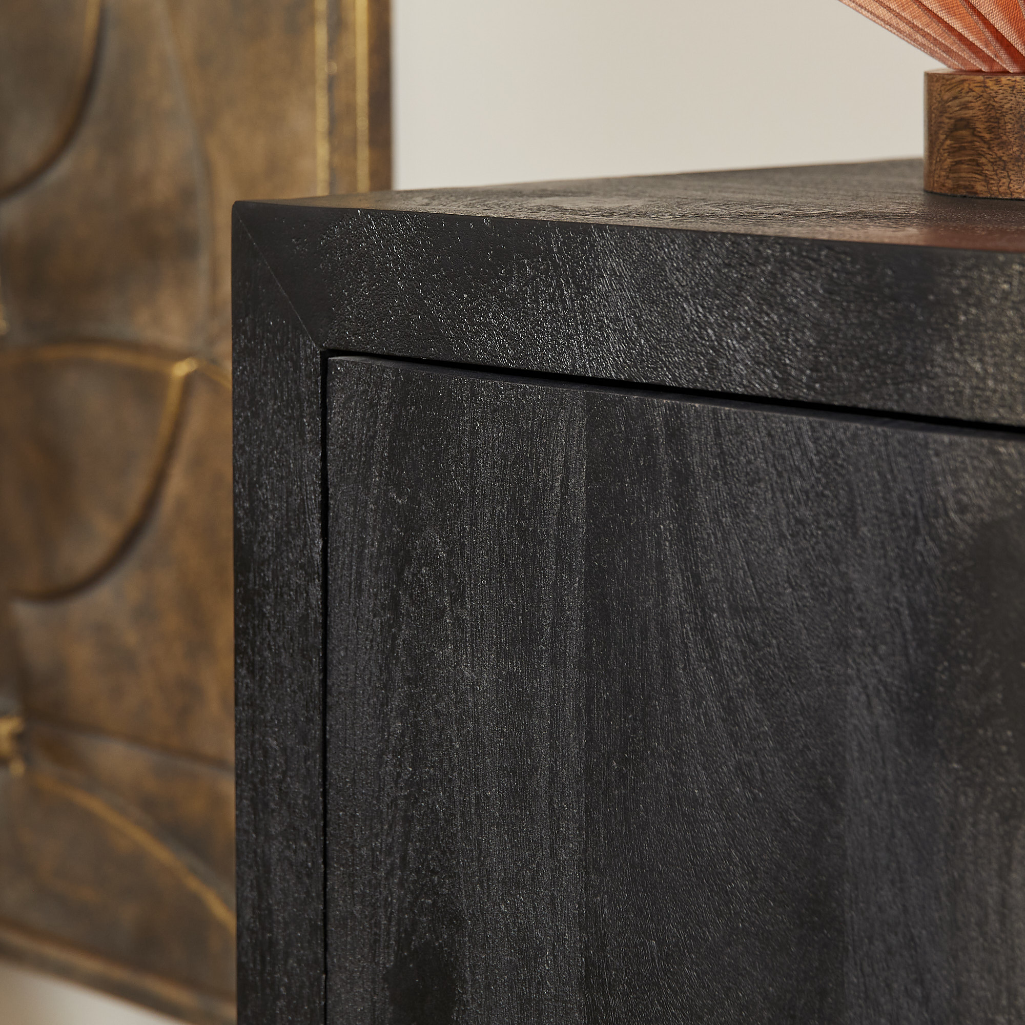 Close-up van de hoek van de Dressoir Brussel Black 3 deurs | 150 cm, met een gladde donkerhouten afwerking. Op de achtergrond zijn ook een deel van een decoratief bronzen wandkunstwerk en een klein roodachtig object bovenop zichtbaar.