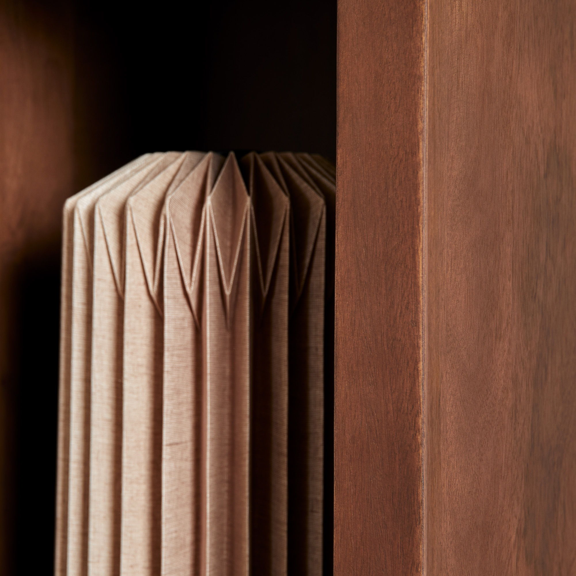 Close-up van de Vakkenkast Brussel Walnut | 90 cm, met het geplooide, geometrische houtpatroon omlijst door gladde verticale walnootpanelen. Verlichting benadrukt de rijke textuur en precieze structuur van het gevouwen materiaal.