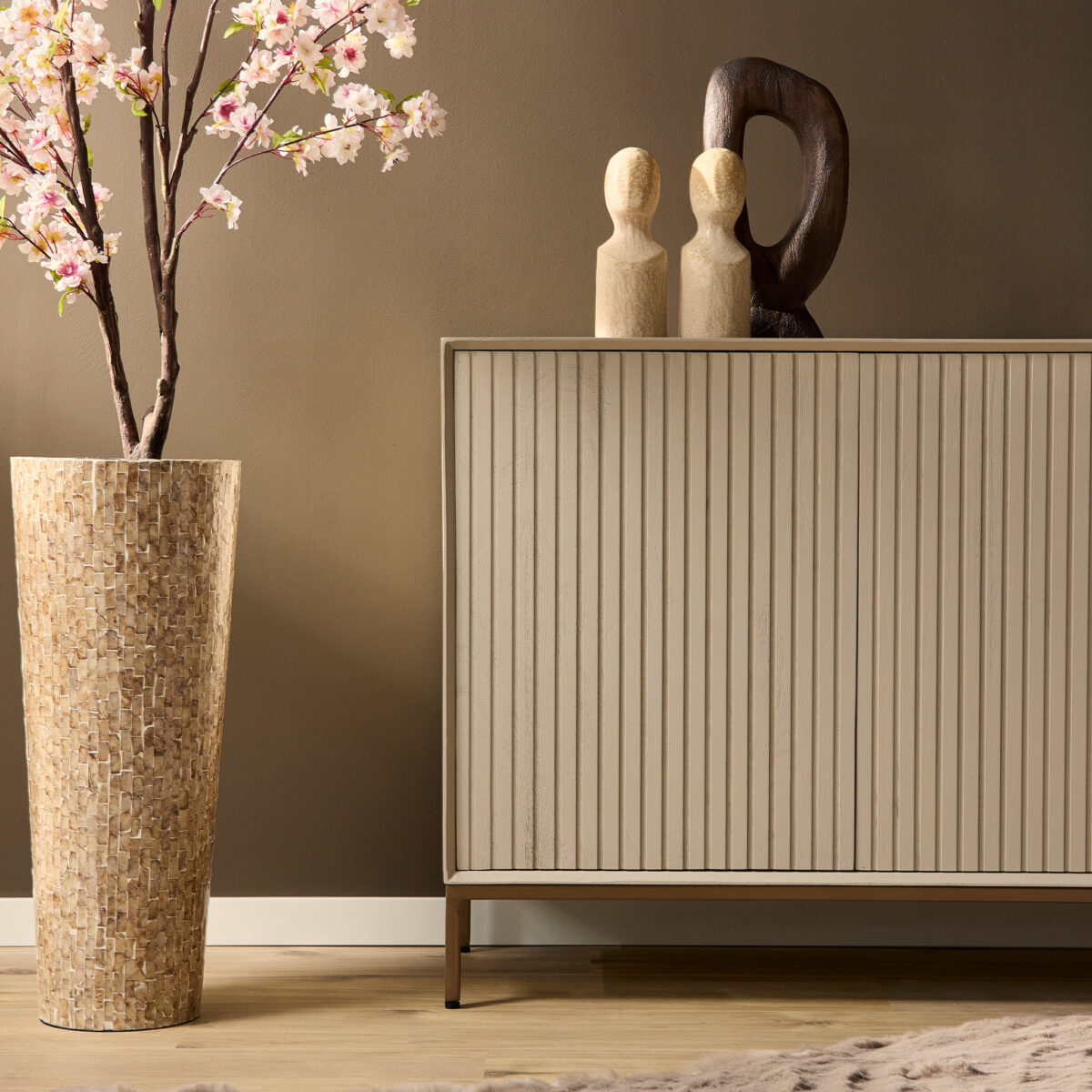 Een hoge mozaïekvaas met roze bloesems staat naast de 210 cm Dressoir Madison Sand, die verticale groeven heeft en plaats biedt aan twee houten beeldjes en een donker abstract beeld, allemaal tegen een taupekleurige muur.