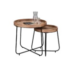 De Mirrors and More Bijzettafel JETTE bestaat uit twee ronde nesttafels met houten bladen en zwarte metalen poten; op de grotere tafel staan een glazen karaf, een drinkglas en een zwart rechthoekig object, terwijl de kleinere er gedeeltelijk onder kan worden genest.