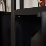 Close-up van de Boekenkast Boaz Black | 1 deur, een zwarte houten boekenplank met metalen steunen. Op de achtergrond verschijnen gedeeltelijk zichtbare objecten en een decoratief voorwerp met een rode top tegen een neutraal kleurenpalet.