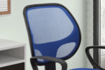 Een close-up van de Bureaustoel 'Hippa' Mesh Blauw (1c), een blauw gazen bureaustoel met een zwart frame, geplaatst bij een bureau en een raam. Op de achtergrond zijn een deel van een witte bakstenen muur en een kurkbord met een kalender zichtbaar.