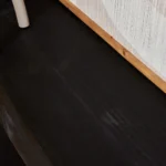 Een close-up van de Sidetable Boaz Black | 120 cm toont het donkere, gestructureerde houten oppervlak met een lichte houten rand en een deel van een beige cilindervormig object in de linkerbovenhoek.