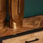 Close-up van de Sidetable Arlington | 2 lades, met een visgraat houten blad, zwarte bies en handvat. Er staan twee grote goudmetalen decorstukken op, tegen een donkergroene muur.