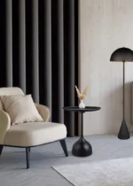 Een moderne woonkamer met een crèmekleurige fauteuil, de Mirrors and More Bijzettafel MONA die een kleine vaas met gedroogde bloemen vasthoudt, een zwarte staande lamp, donkere gordijnen en lichthouten muren en vloeren.
