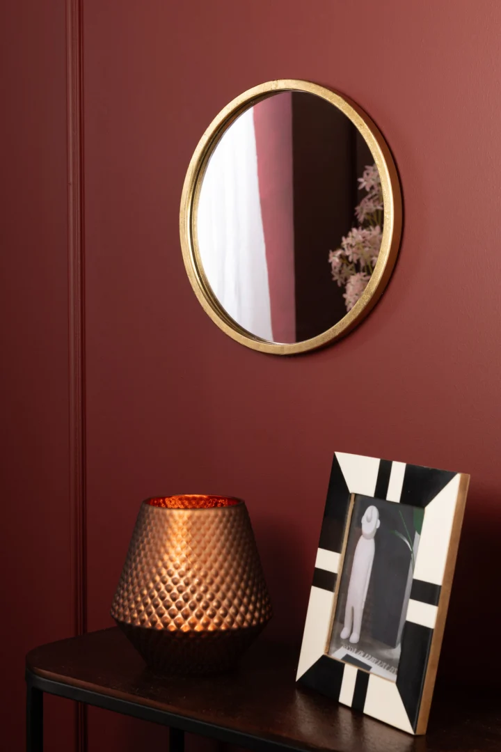 Een extra kleine ronde Mona spiegel met een gouden frame van ijzer en glas hangt aan een rode muur boven een donkere tafel. Op de tafel staan een koperen kandelaar met textuur en een zwart-wit fotolijstje met abstracte kunst.