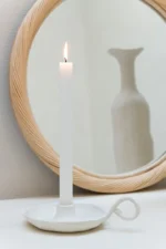 Een brandende witte kaars in een eenvoudige houder staat voor de wandspiegel Round Rattan/Glass Natural Medium, die de kaars en een vage witte vaas op de achtergrond weerspiegelt.