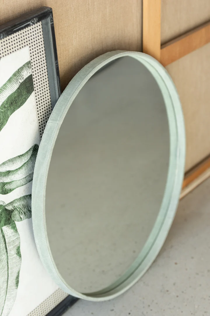 Een kleine ronde spiegel met een groene leren lijst leunt tegen een beige muur, naast een ingelijst kunstwerk van groene bladeren. De licht gespikkelde vloer versterkt de look.