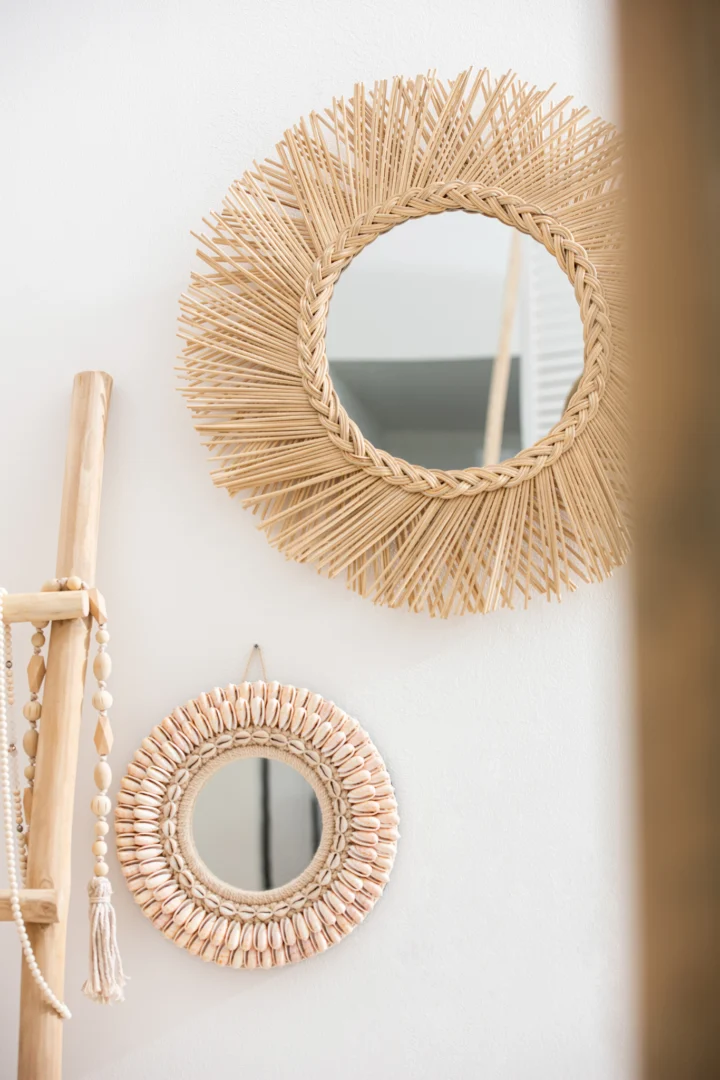 Twee Mirror Tress Rattan Natural-elementen met geweven frames van natuurlijke vezels - een zonnestraal en een opgerold ontwerp - hangen aan een witte muur, terwijl een houten ladder met kralen vlakbij leunt.