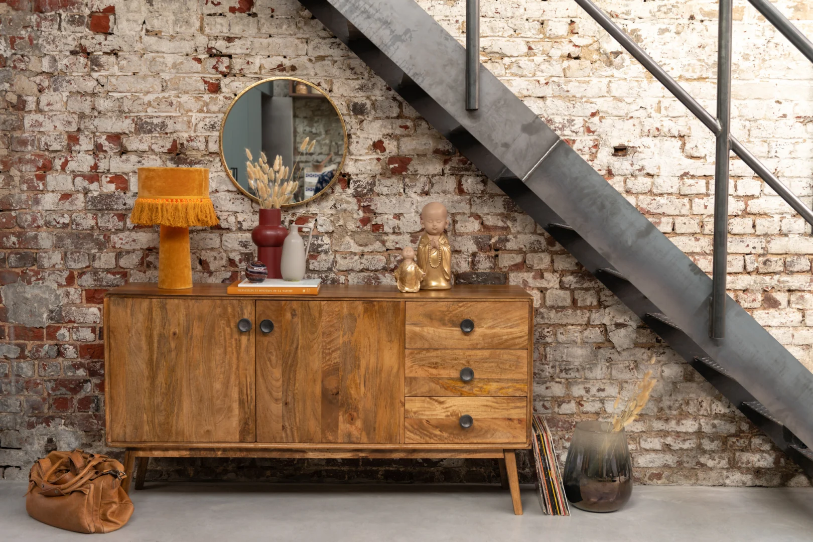 Een houten dressoir staat tegen een witte bakstenen muur onder een metalen trap, met daarop de Mirror Mona Round Iron/Glass Gold Large, een lamp, vazen, decoratieve voorwerpen en boeken. Een bruine tas en een vaas met gedroogd gras staan op de vloer vlakbij.