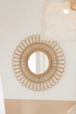 De Spiegel Ellen Round Rattan Natural Small met een decoratief sunburst frame hangt aan een wit-beige muur; een deel van een geweven lampenkap verschijnt in de rechterbovenhoek.