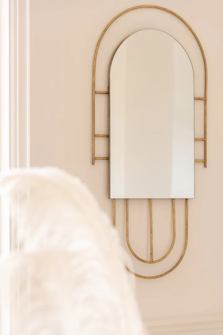 De spiegel Ines Metal Gold, met een modern geometrisch ontwerp met een gebogen bovenkant en rechthoekige onderkant, hangt aan een lichte muur. Het goudkleurige metalen frame is abstract geaccentueerd, terwijl wit pampagras zacht vervaagt op de voorgrond.