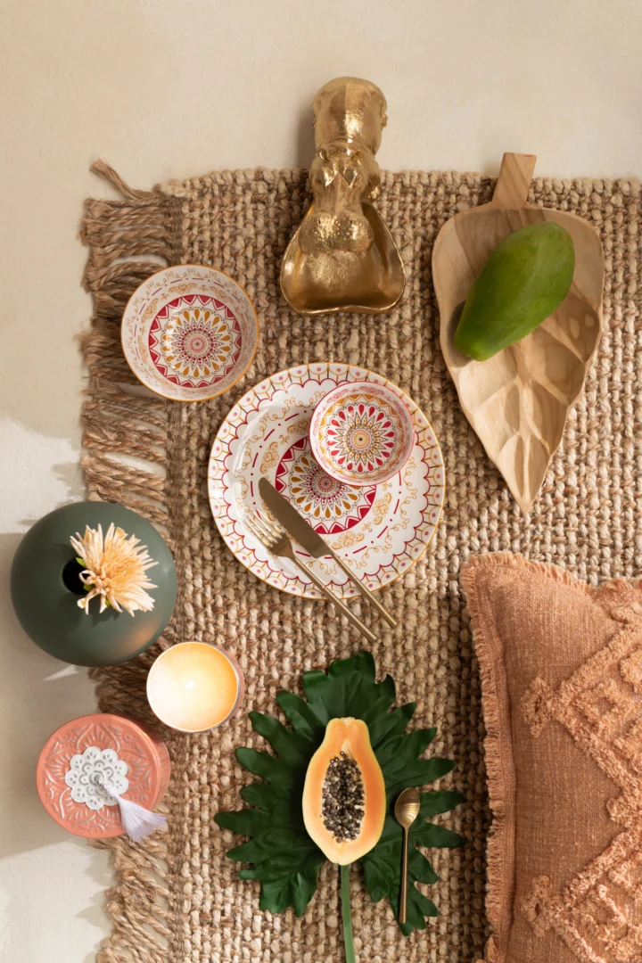 Een flatlay toont gedessineerde borden, gouden bestek, een bloem in een vaas, een gouden nijlpaardbeeldje, papaja, mango en kaarsen gerangschikt op het Vloerkleed Havana Poly Naturel/Wit Klein vloerkleed met een rand met franjes en een kussen met oranje structuur.