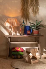 Een houten consoletafel met een rotan lamp, kleurrijke beeldjes, een potplant en decoratieve borden. Eronder liggen lichte kussens en op het Vloerkleed Rond Gevlochten Zeegras Naturel ligt een gebeeldhouwde houten kruk. Gedroogd gras siert de muur.