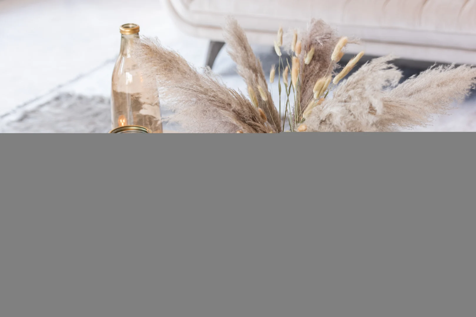 Een heldere glazen fles en een boeket van gedroogd pampagras en gebladerte zijn gerangschikt op een tafel, met een beige bank en het Vloerkleed Langharig Acryl Grijs zachte vloerkleed op de achtergrond.