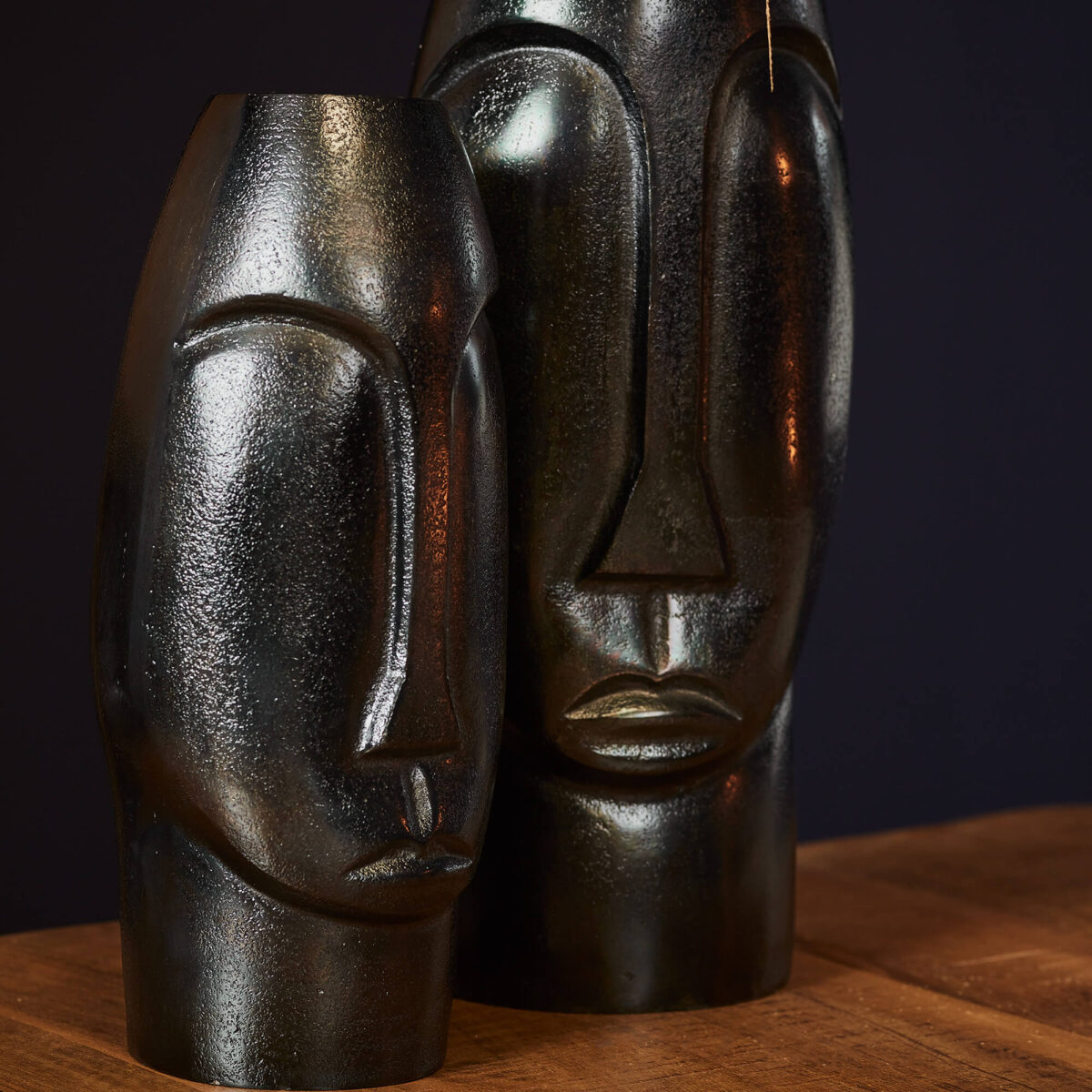 Twee grote, zwarte Vaas Lyra - Zwart sculpturen met gestileerde gezichten en gladde oppervlakken staan naast elkaar op een houten tafel tegen een donkere achtergrond.