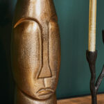 Een close-up van de Vaas Lyra - Antiek goud | Large, een metallic gouden abstract gezichtssculptuur, uitgestald naast een zwarte kandelaar met een hoge kaars op een houten oppervlak tegen een groene muur.