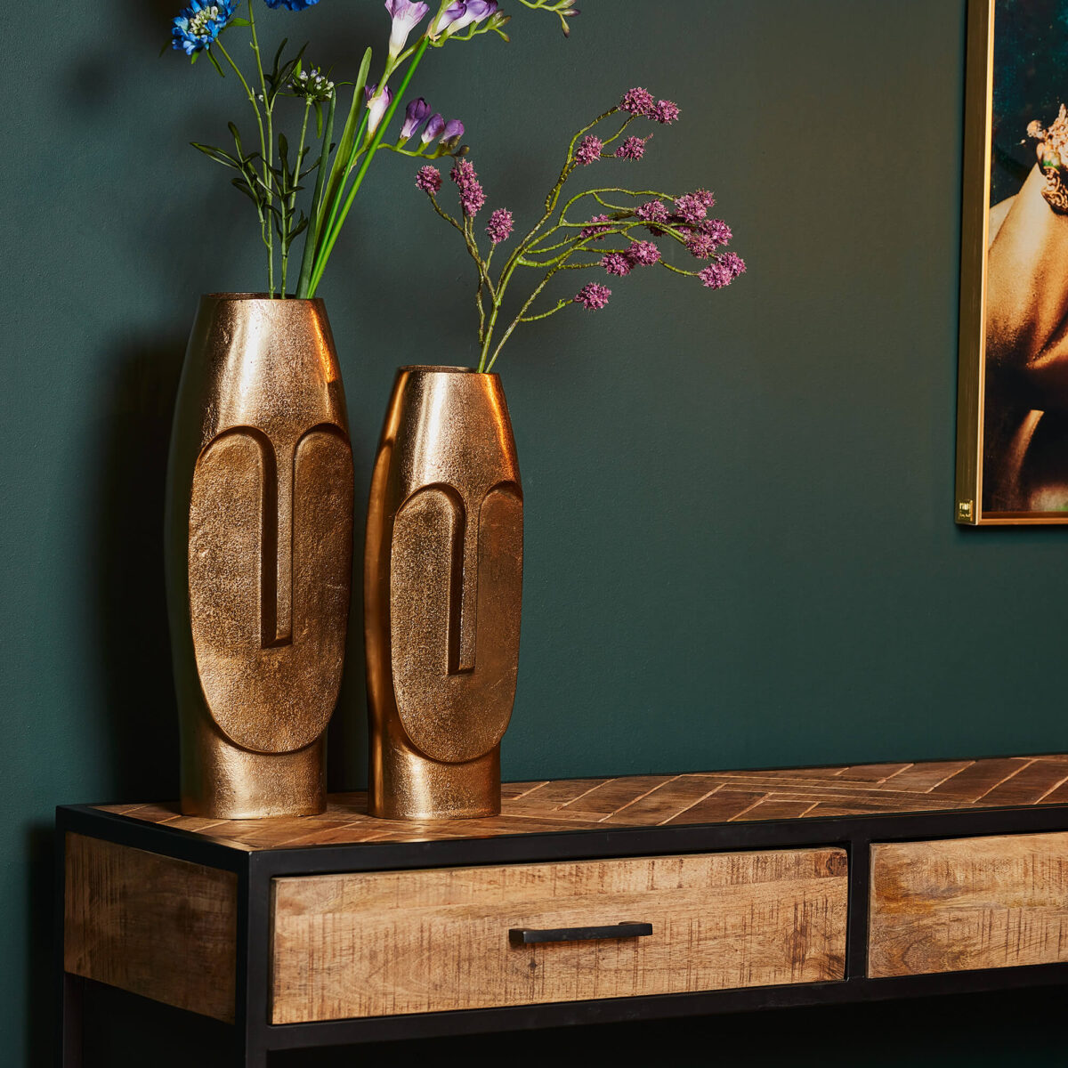 Twee hoge Vaas Liv - Antiek goud | Grote abstracte gezichtsvazen met bloemen staan op een houten tafel met zwart metalen poten en laden, tegen een donkergroene muur met rechts een ingelijst kunstwerk.